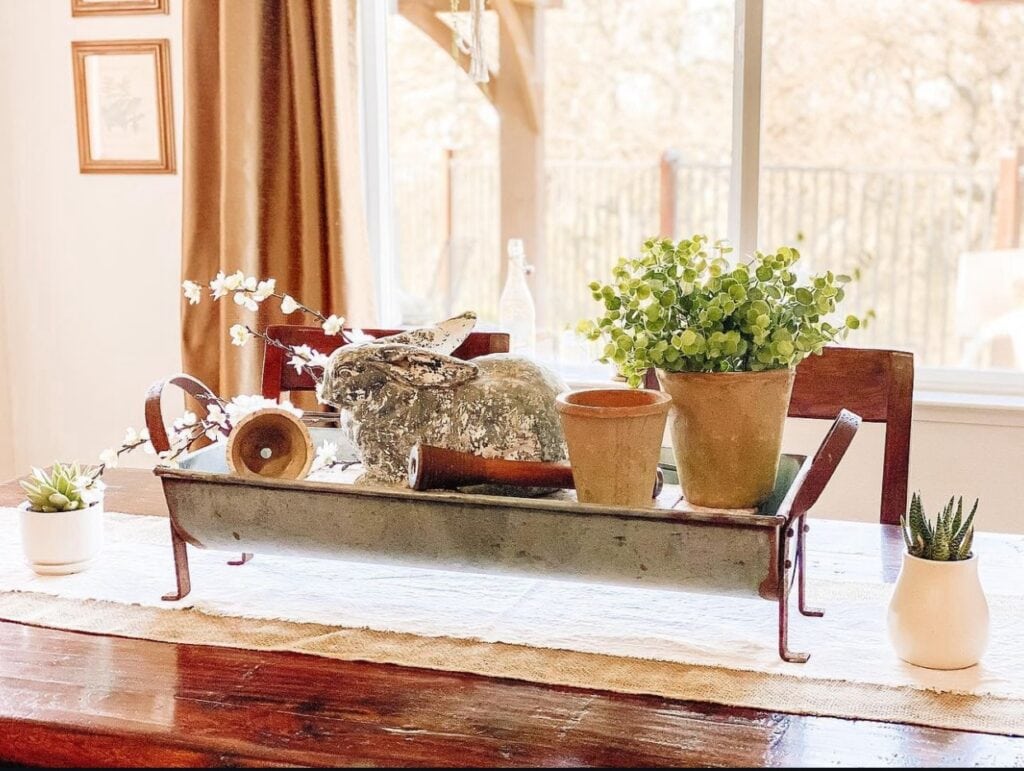 An Easter Centerpiece on a dining room table with a bunny figurine and terra cotta pots with faux flowers and plants